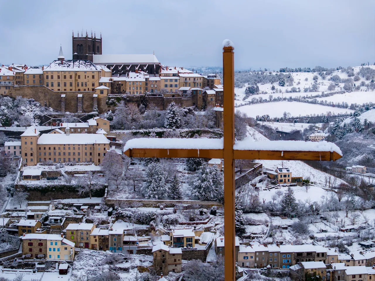 Ville de Saint-Flour enneigée vue depuis les hauteurs, avec une grande croix en bois au premier plan